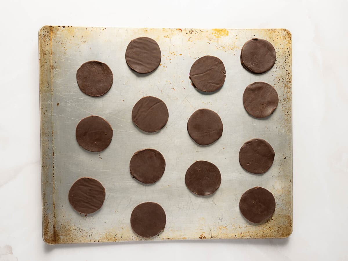 A metal baking sheet with 15 evenly spaced, round chocolate cookie dough discs arranged in rows, ready to be baked.