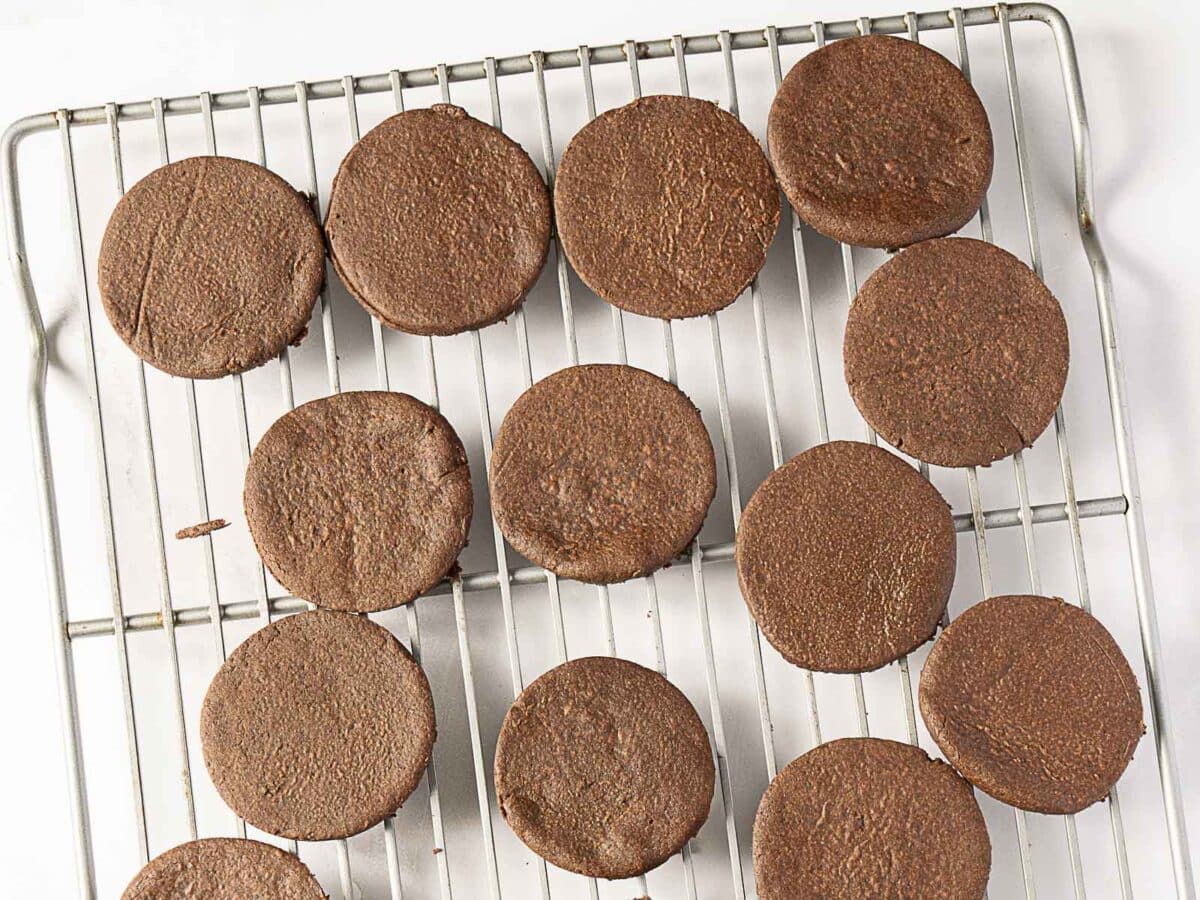 Round chocolate cookies cooling on a metal wire rack, with a white background visible underneath.