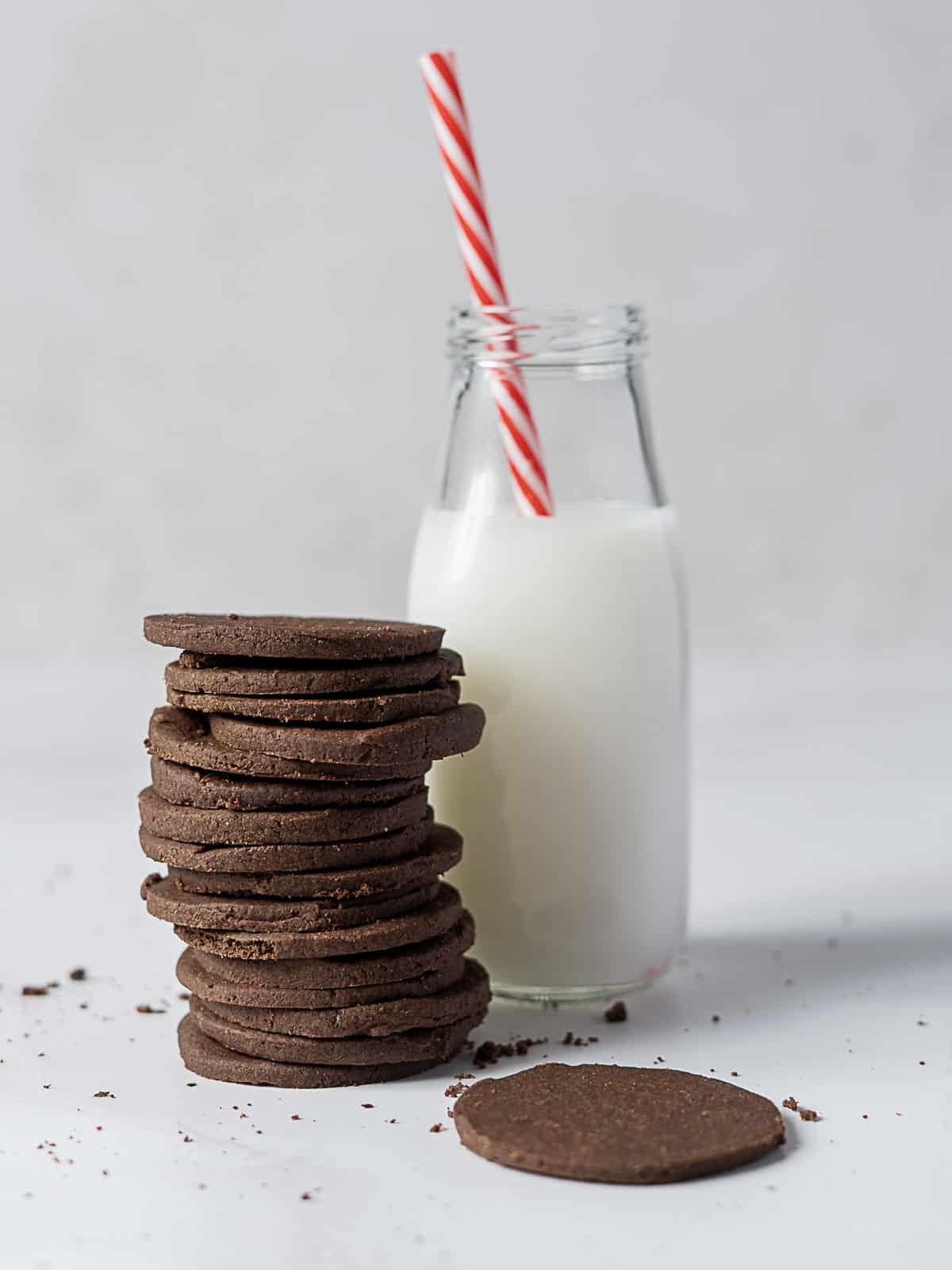 A stack of chocolate cookies sits next to a glass bottle of milk with a red and white striped straw; cookie crumbs are scattered on the surface.