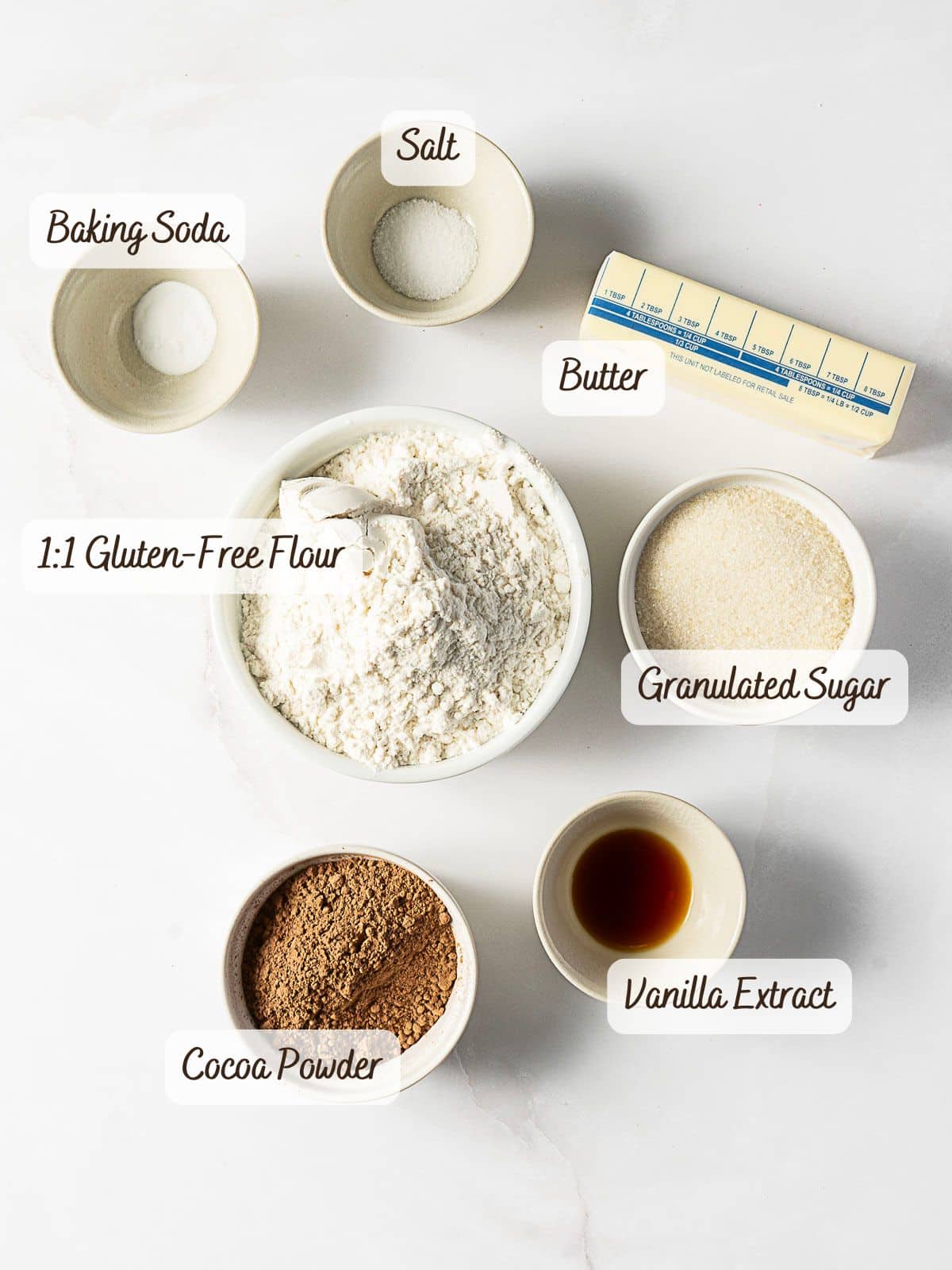 Seven baking ingredients for Gluten Free Chocolate Wafer Cookies-baking soda, salt, butter, 1:1 gluten-free flour, granulated sugar, cocoa powder, and vanilla extract-are arranged in bowls on a white surface.