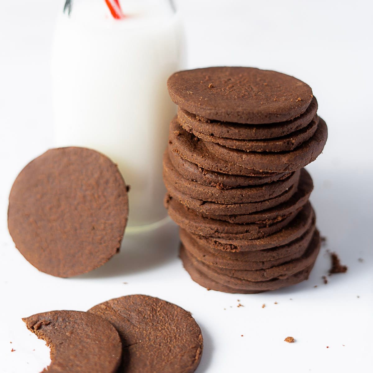 A stack of round chocolate cookies next to a glass bottle of milk, with two cookies lying flat in front, one with a bite taken out.
