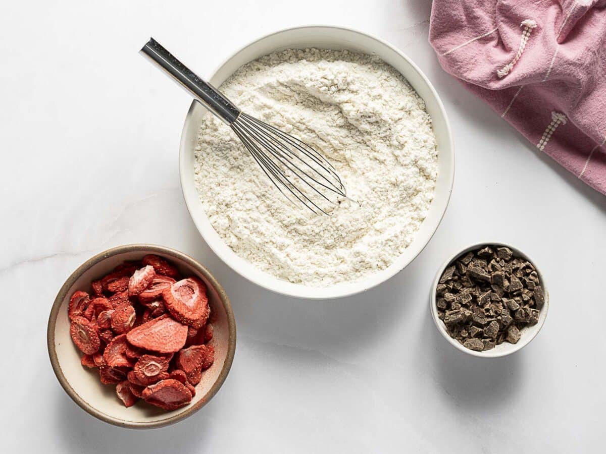 A bowl of flour with a whisk, a bowl of freeze-dried strawberries, and a bowl of chocolate chunks on a white surface with a pink cloth.