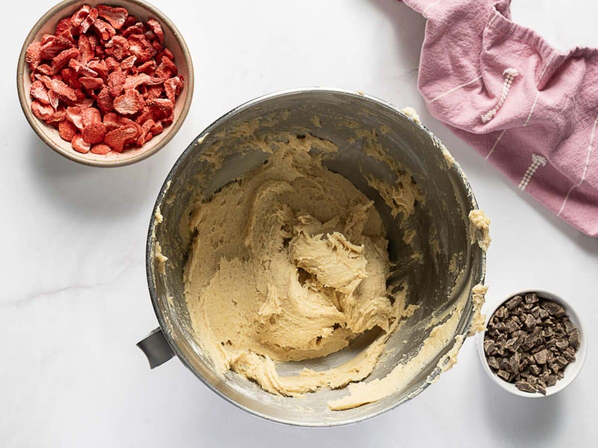 A mixing bowl with cookie dough, a bowl of freeze-dried strawberries, and a cup of chocolate chunks on a white surface with a pink cloth nearby.