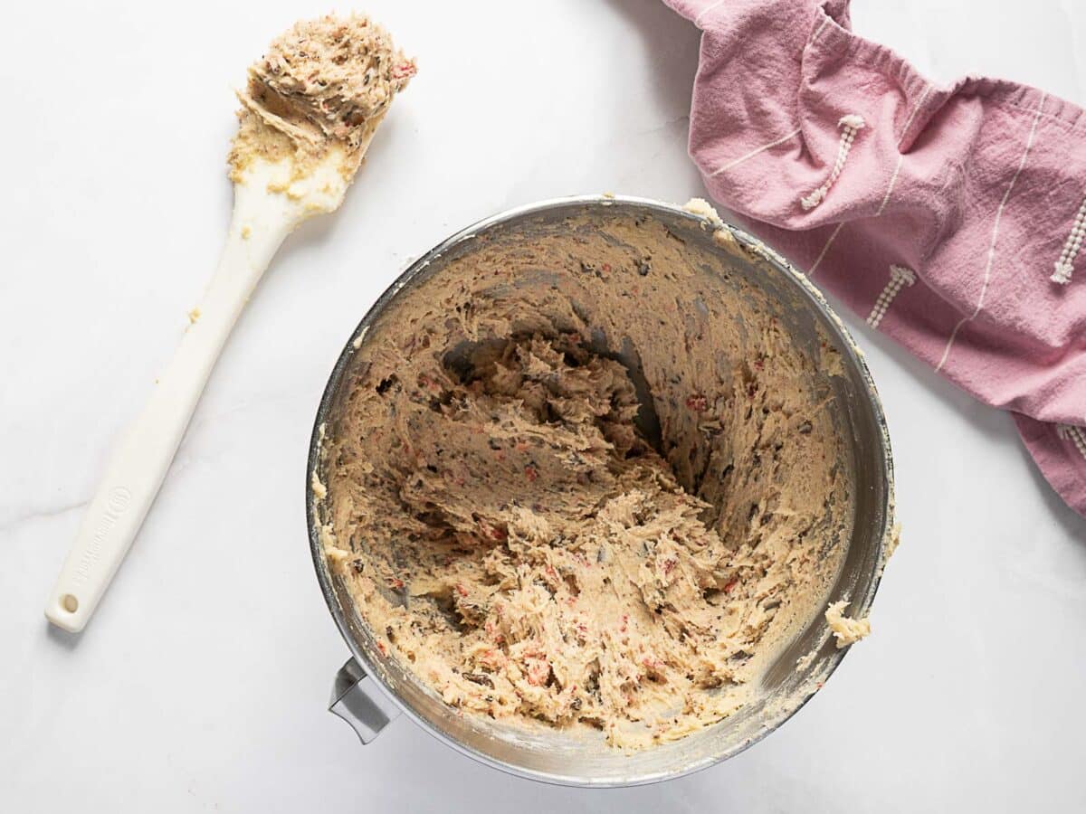 A metal mixing bowl filled with cookie dough sits on a white surface next to a spatula with dough and a folded pink kitchen towel.
