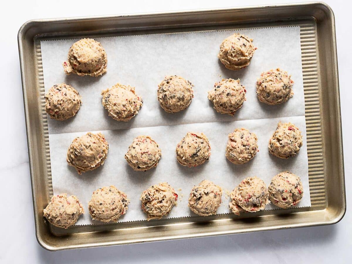 A baking tray lined with parchment paper holds 18 evenly spaced scoops of raw cookie dough.
