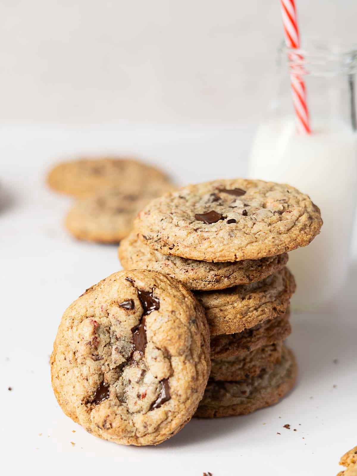 A stack of chocolate chip cookies sits next to a glass bottle of milk with a red and white striped straw on a white surface.