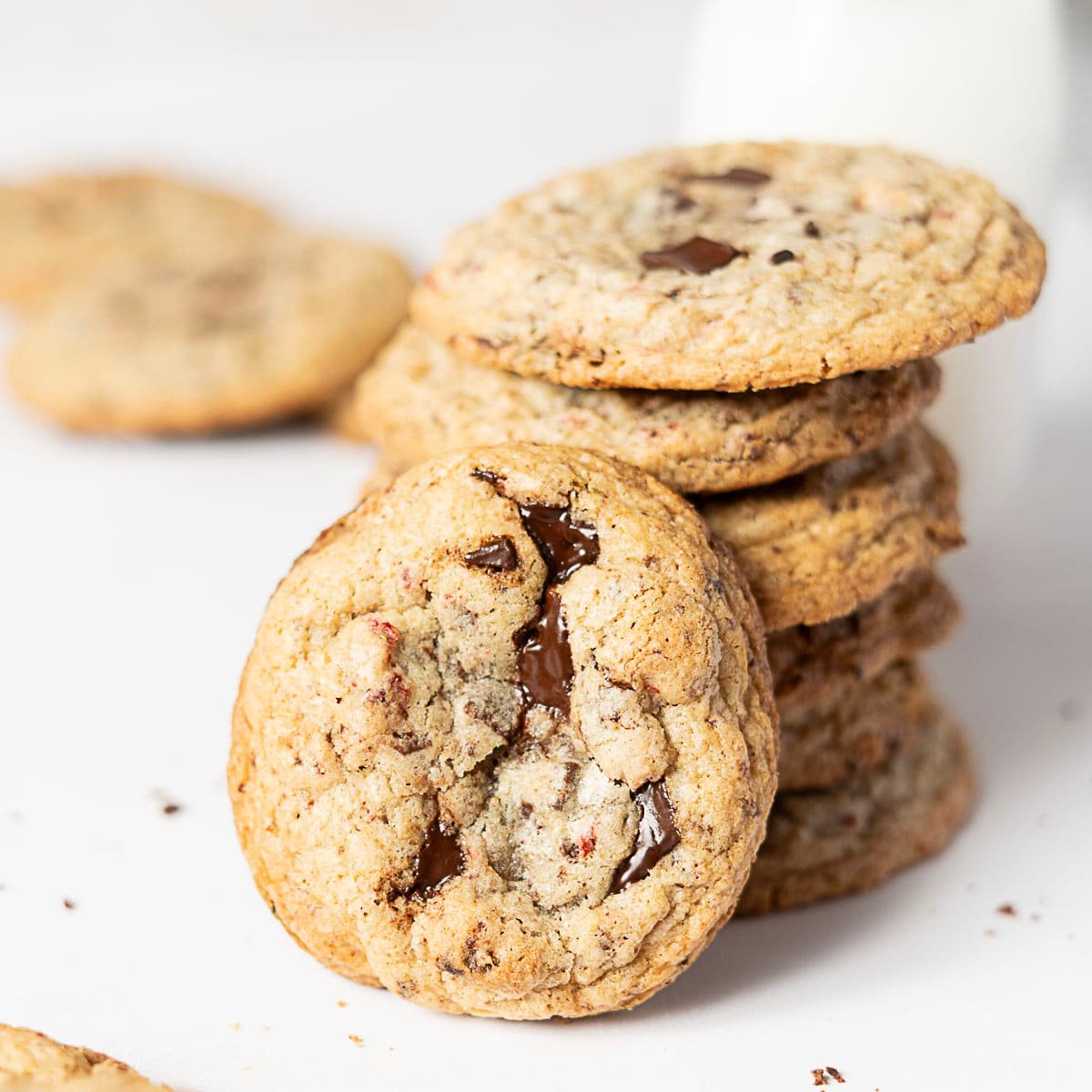 A stack of chocolate chip cookies with one cookie leaning in front, showing melted chocolate pieces; a glass of milk is blurred in the background.