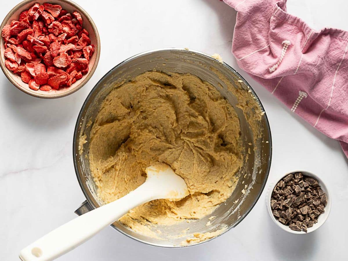 A mixing bowl with cookie dough and a spatula, surrounded by a bowl of dried strawberries, a bowl of chocolate chunks, and a pink kitchen towel.