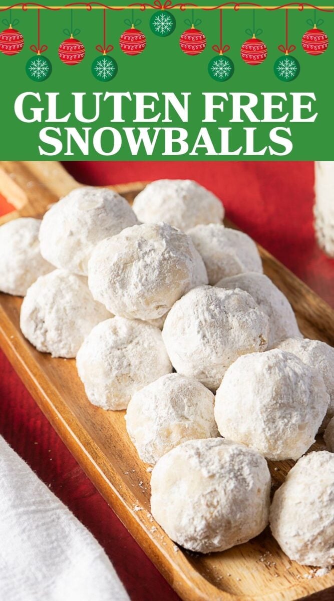 A wooden tray holds a pile of round, powdered sugar-coated gluten free snowball cookies on a red tablecloth.