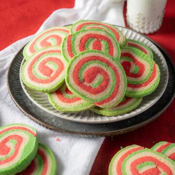 A plate of spiral cookies with red, green, and white swirls arranged on a white cloth, with a glass of milk in the background.