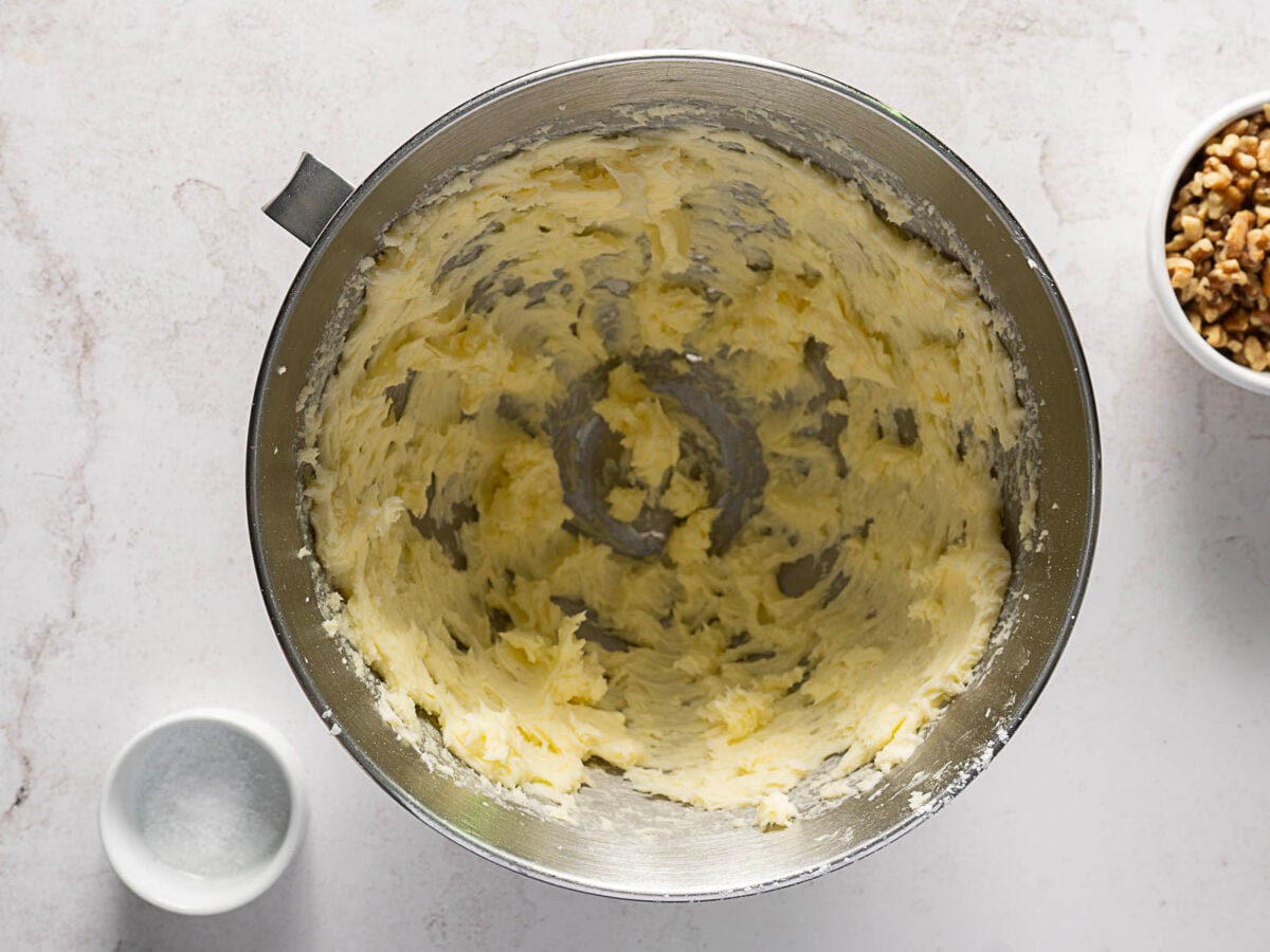 A metal mixing bowl with creamed butter and sugar sits on a white surface, next to a small bowl of salt and a bowl of chopped walnuts.