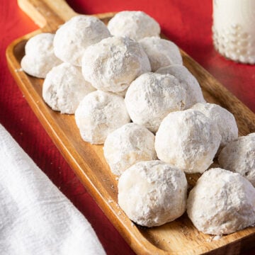 A wooden tray holds several round cookies coated in powdered sugar, set on a red surface next to a glass of milk and a white napkin.