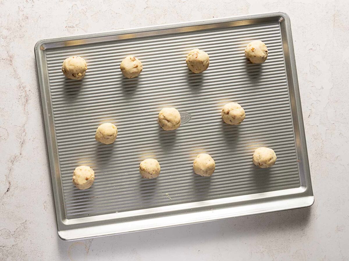 A metal baking sheet with 11 evenly spaced balls of cookie dough, ready to be baked, on a light-colored surface.