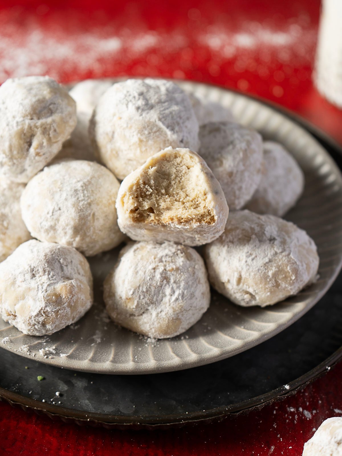 A plate of round, powdered sugar-coated cookies, with one cookie showing a bite taken out to reveal its crumbly interior, set against a red background.