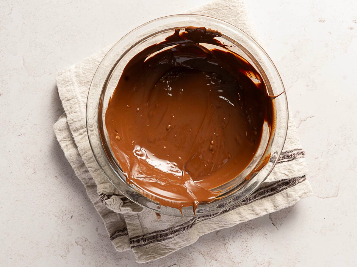 A glass bowl filled with melted chocolate, ready to become a luscious no bake chocolate cake, sits on a folded kitchen towel on a light-colored surface.