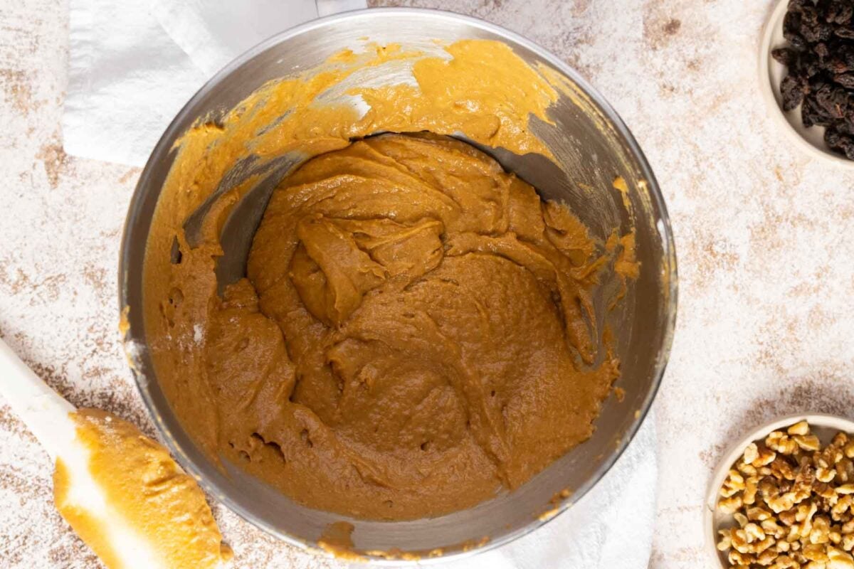 A metal mixing bowl filled with thick, light brown Gluten Free Dairy Free Carrot Cake batter sits on a countertop, surrounded by a spatula, walnuts, and raisins.