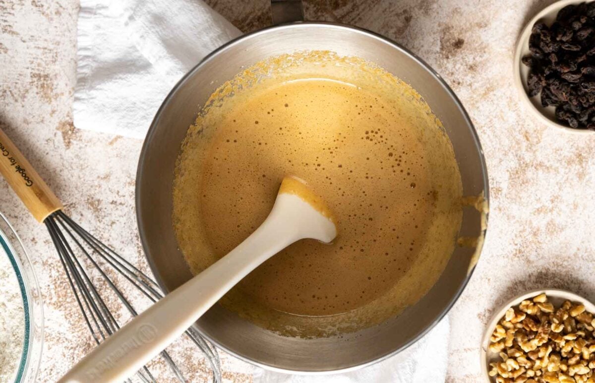 Mixing bowl with light brown Gluten Free Dairy Free Carrot Cake batter and a spatula inside, surrounded by a whisk, raisins, and chopped walnuts on a light countertop.