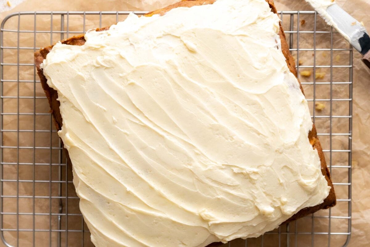 A Gluten Free Dairy Free Carrot Cake with white frosting spread on top sits on a wire cooling rack over parchment paper.