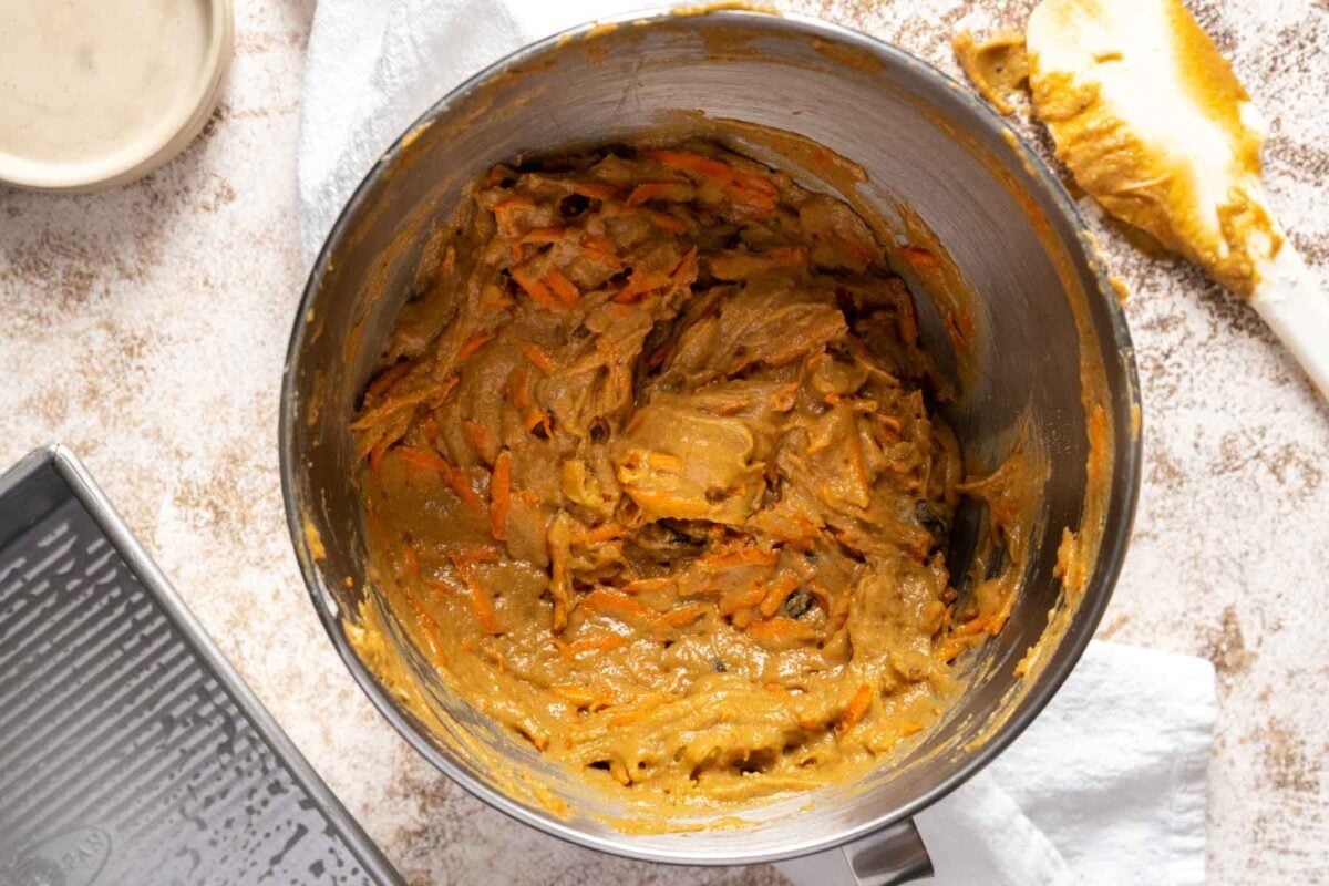 A mixing bowl filled with gluten free dairy free carrot cake batter, with visible grated carrots, sits on a countertop next to a spatula and a baking pan.