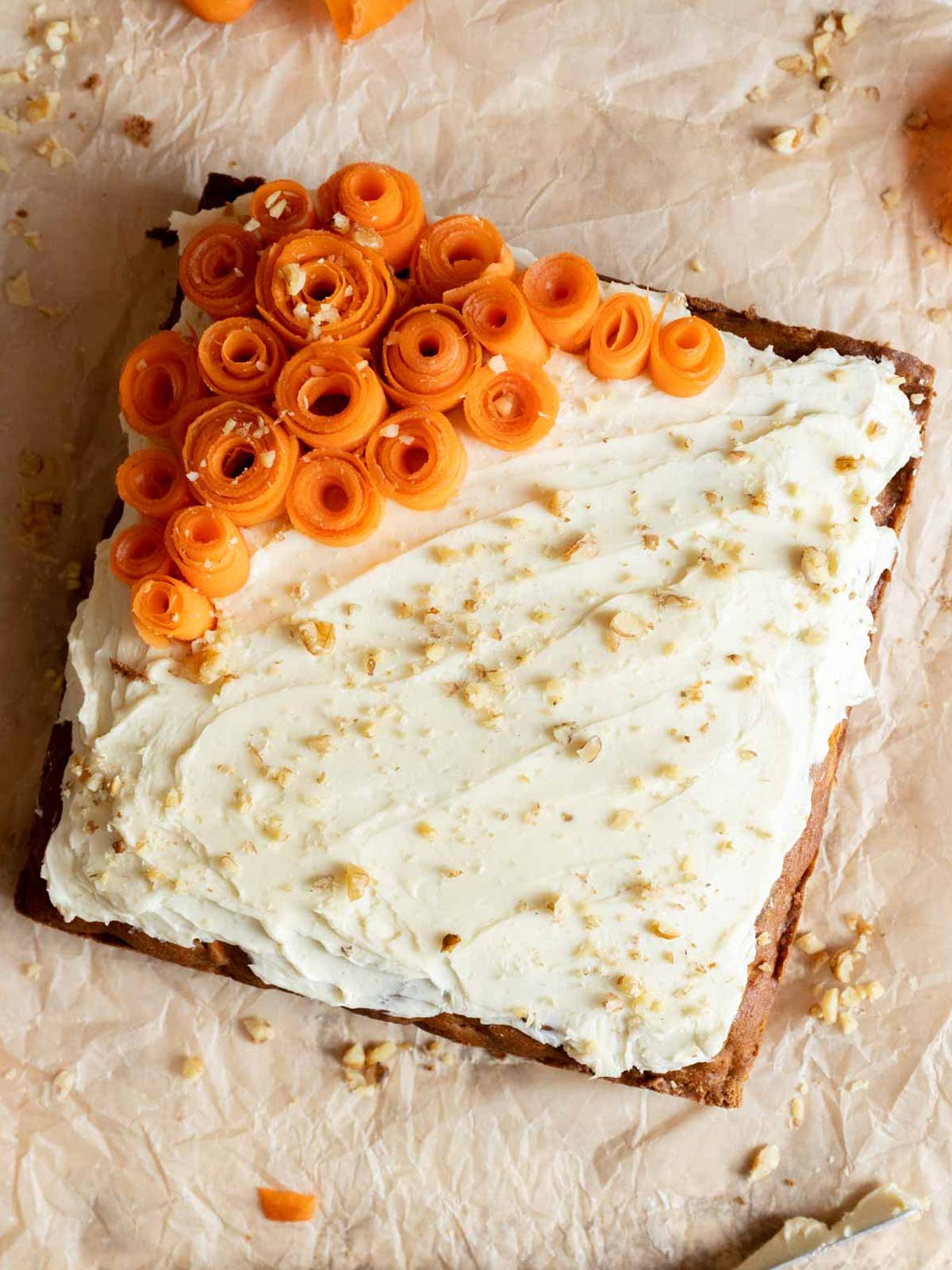 A square Gluten Free Dairy Free Carrot Cake with white frosting is topped with curled carrot ribbons arranged in one corner and sprinkled with crushed nuts.