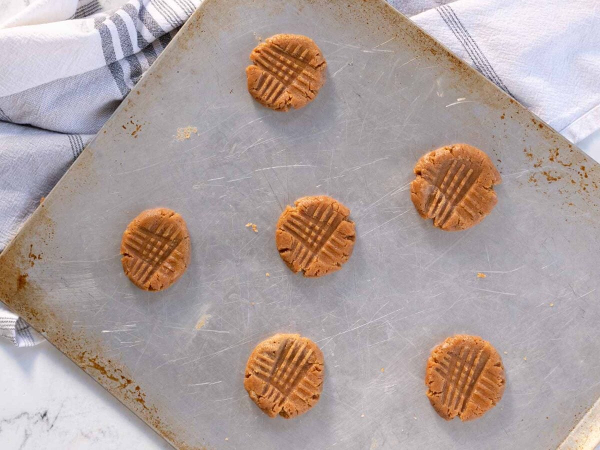 Six unbaked peanut butter cookies arranged on a metal baking sheet, with a kitchen towel partially visible in the background.