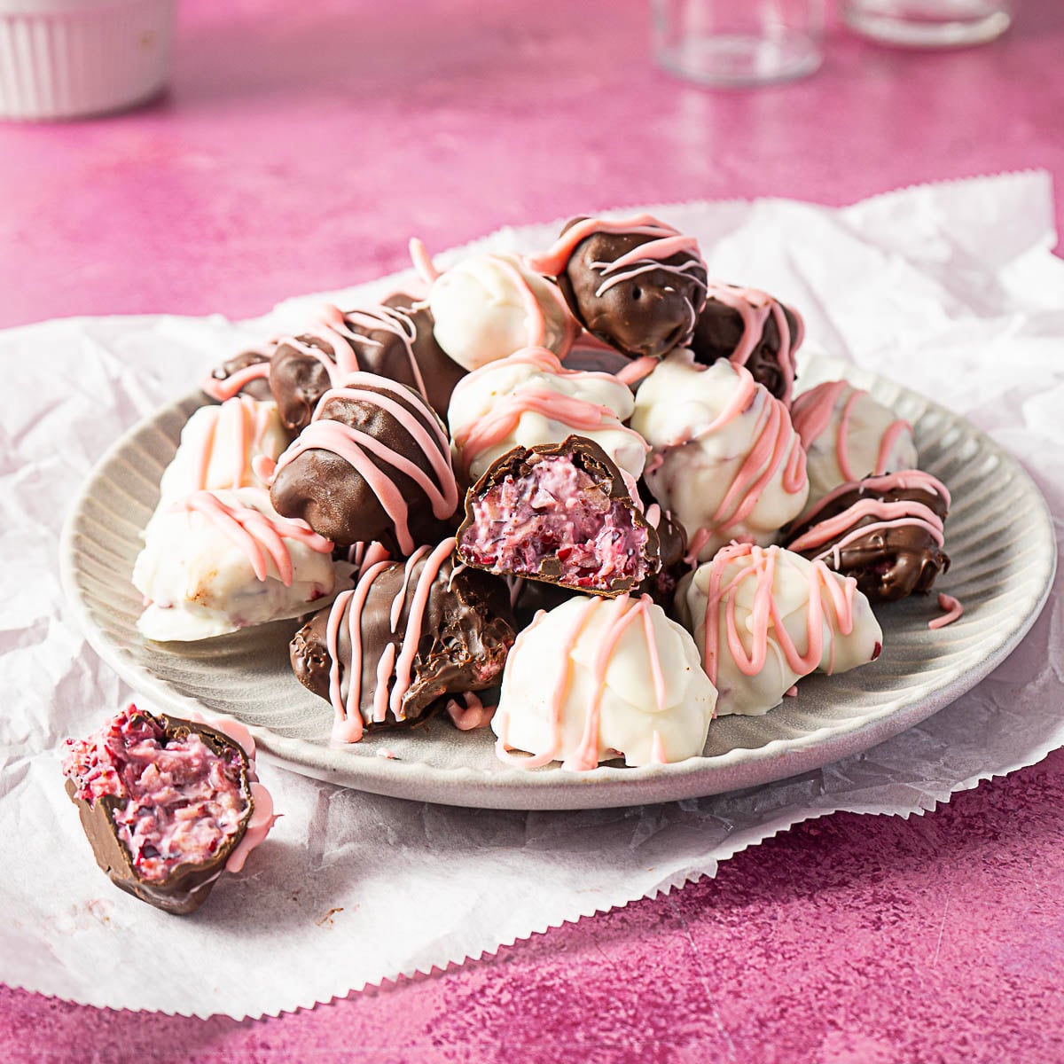 A plate of fresh chocolate bites and white chocolate coated truffles with pink drizzle, some revealing a cranberry pink creamy filling, on white parchment paper against a pink background.