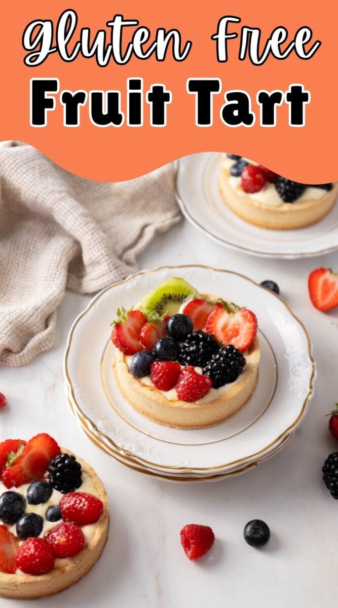 Three gluten-free fruit tarts topped with strawberries, blueberries, raspberries, blackberries, and kiwi are served on white plates with a beige cloth nearby.