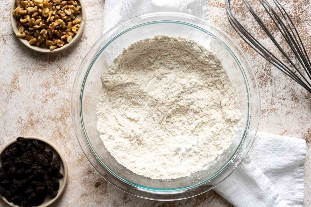 A glass bowl filled with flour mixture sits on a countertop, surrounded by a whisk, a white towel, a bowl of walnuts, and a bowl of raisins-perfect prep for Gluten Free Dairy Free Carrot Cake.