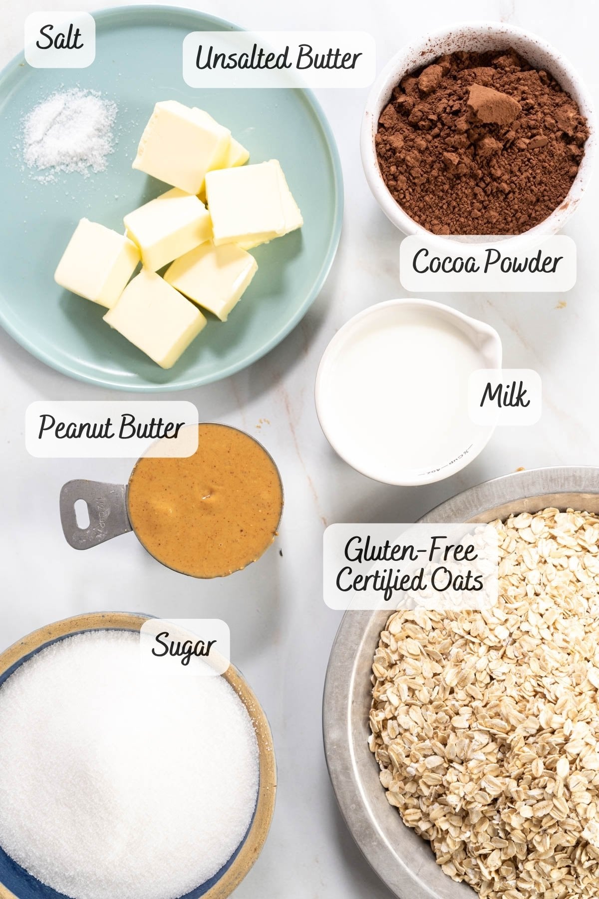 Various baking ingredients, including salt, unsalted butter, cocoa powder, milk, peanut butter, gluten-free certified oats, and sugar, arranged in separate bowls on a white surface-perfect for making Gluten-Free No Bake Cookies.