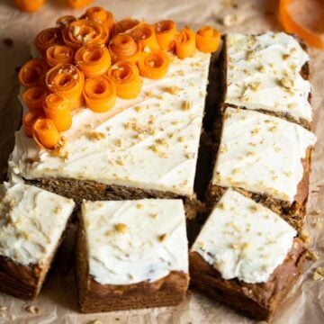 A rectangular Gluten Free Dairy Free Carrot Cake with white frosting, topped with carrot ribbons and chopped nuts, partially sliced into squares on parchment paper.