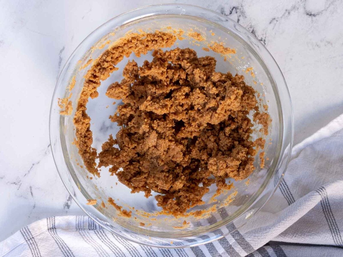 A glass bowl filled with mixed brown cookie dough sits on a marble surface next to a folded white and gray striped kitchen towel.