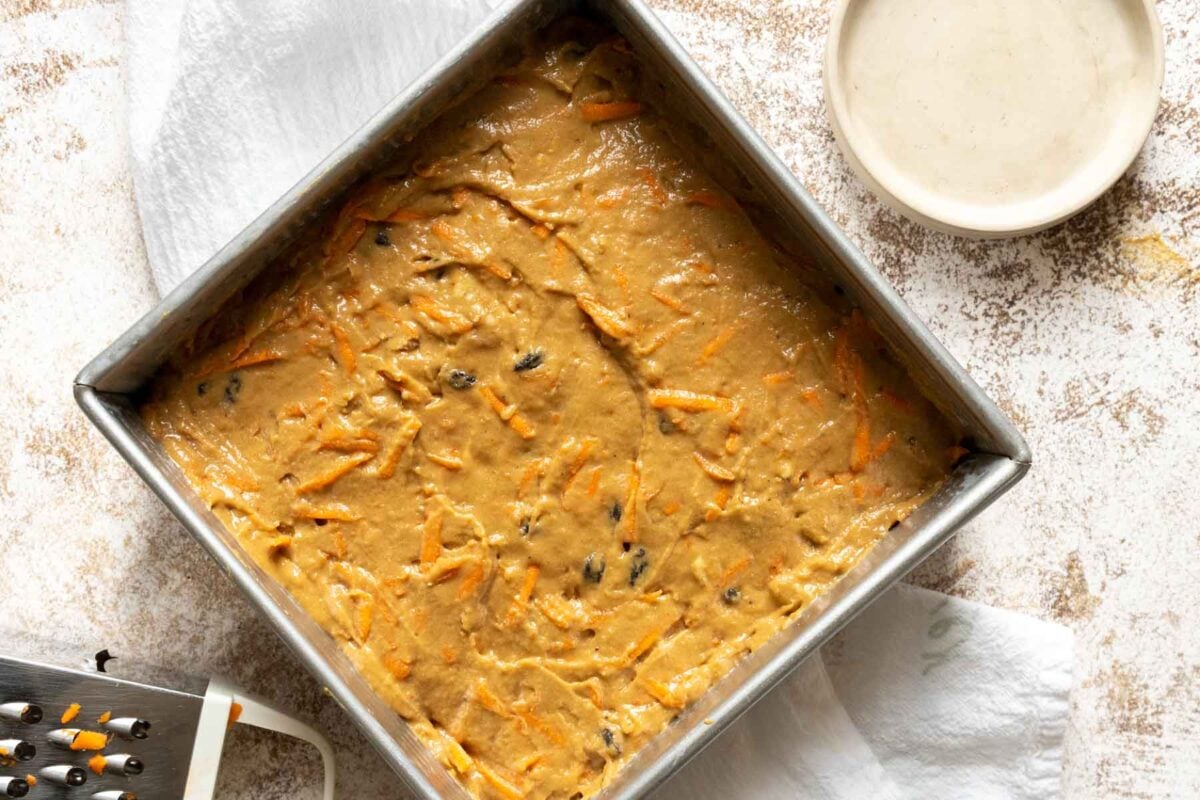 Square pan filled with unbaked Gluten Free Dairy Free Carrot Cake batter, with visible shredded carrots and raisins, sits on a white towel next to a bowl of frosting and a grater.