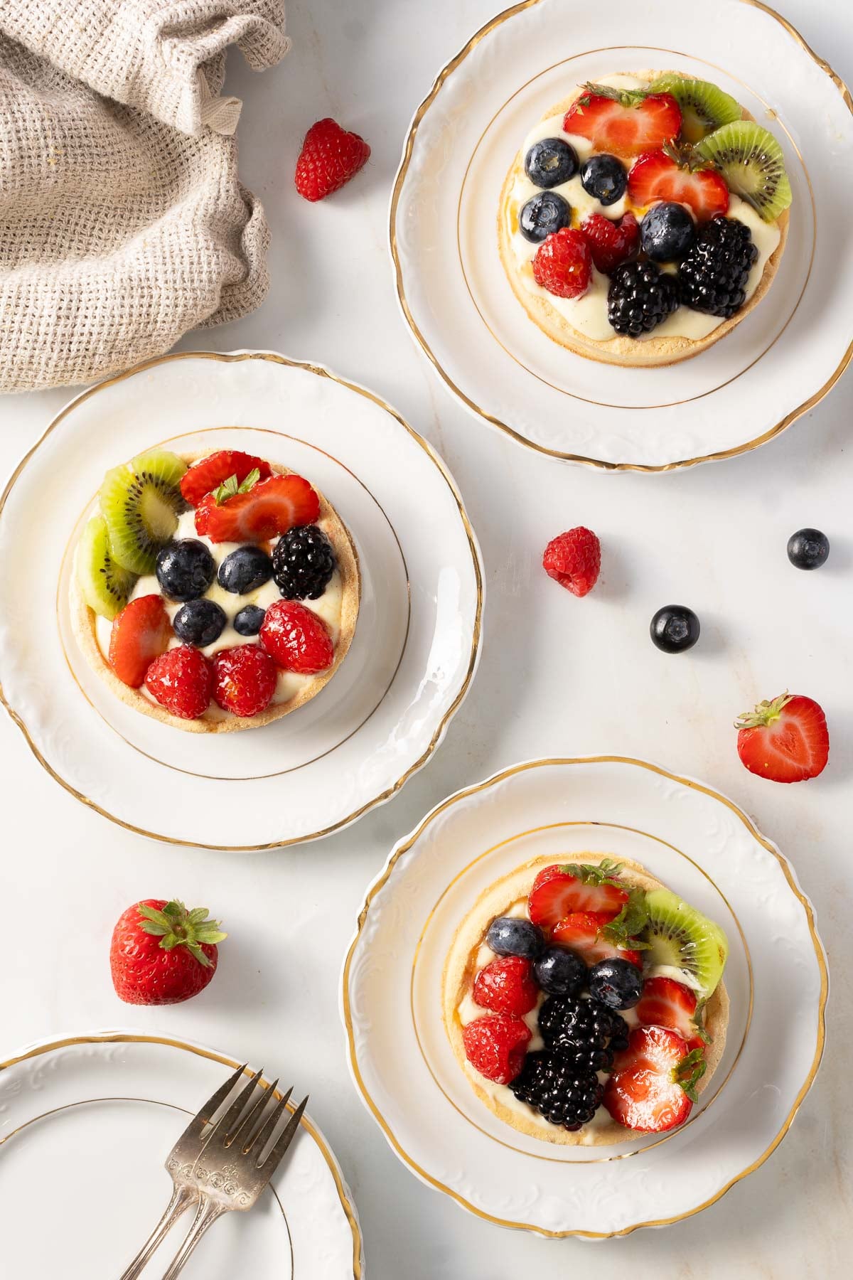 Three small fruit tarts topped with strawberries, blueberries, blackberries, and kiwi are served on white plates, surrounded by loose fruit, a fork, and a beige napkin on a marble surface.