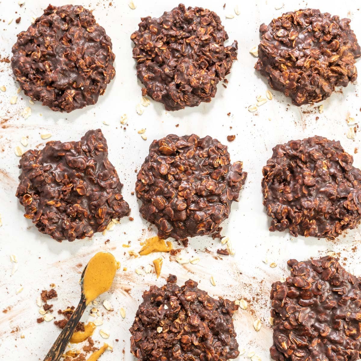 Eight chocolate oat cookies are arranged on a white surface with a spoonful of peanut butter nearby and scattered oats and cocoa powder around them.