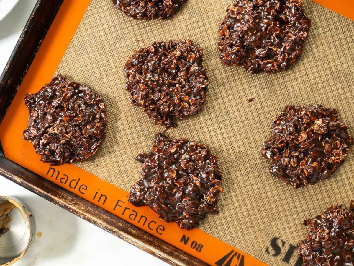 Six gluten-free cookies rest on a silicone baking mat atop a baking sheet; a metal cookie cutter is visible in the corner.