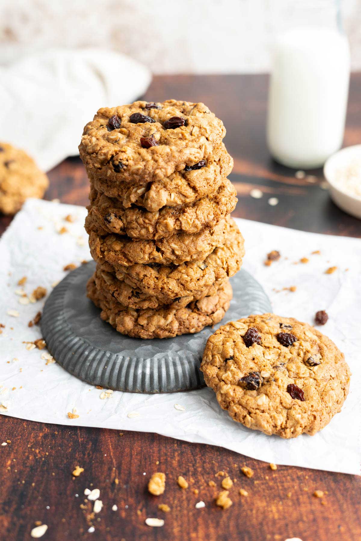 A stack of oatmeal raisin cookies on a metal plate with one cookie leaning against the stack; a glass of milk is in the background.
