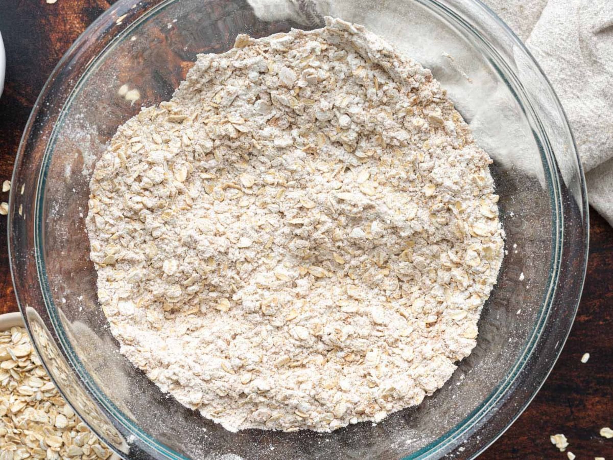 A clear glass bowl filled with a dry mixture of oats and flour sits on a dark wooden surface.