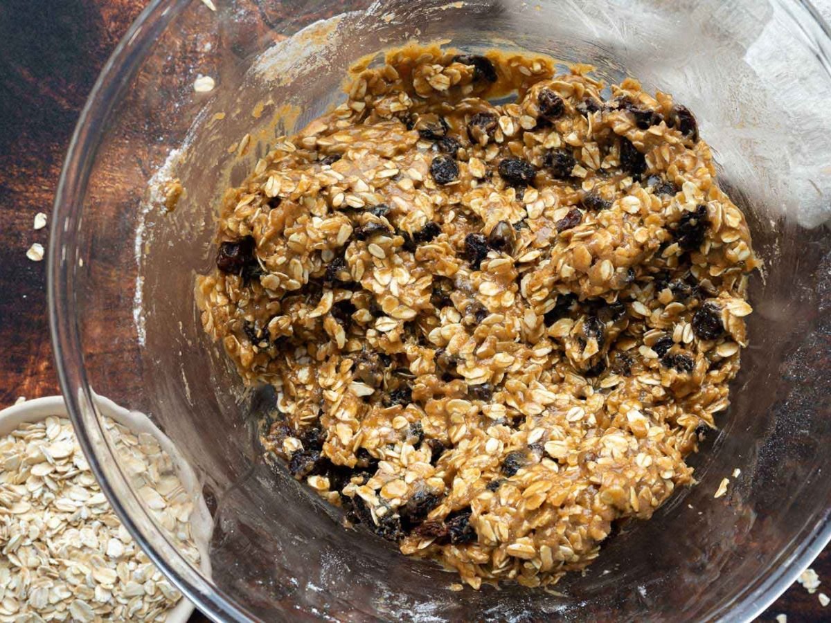 A glass bowl containing oatmeal cookie dough with oats and raisins mixed in. A small bowl of dry oats is partially visible beside it.