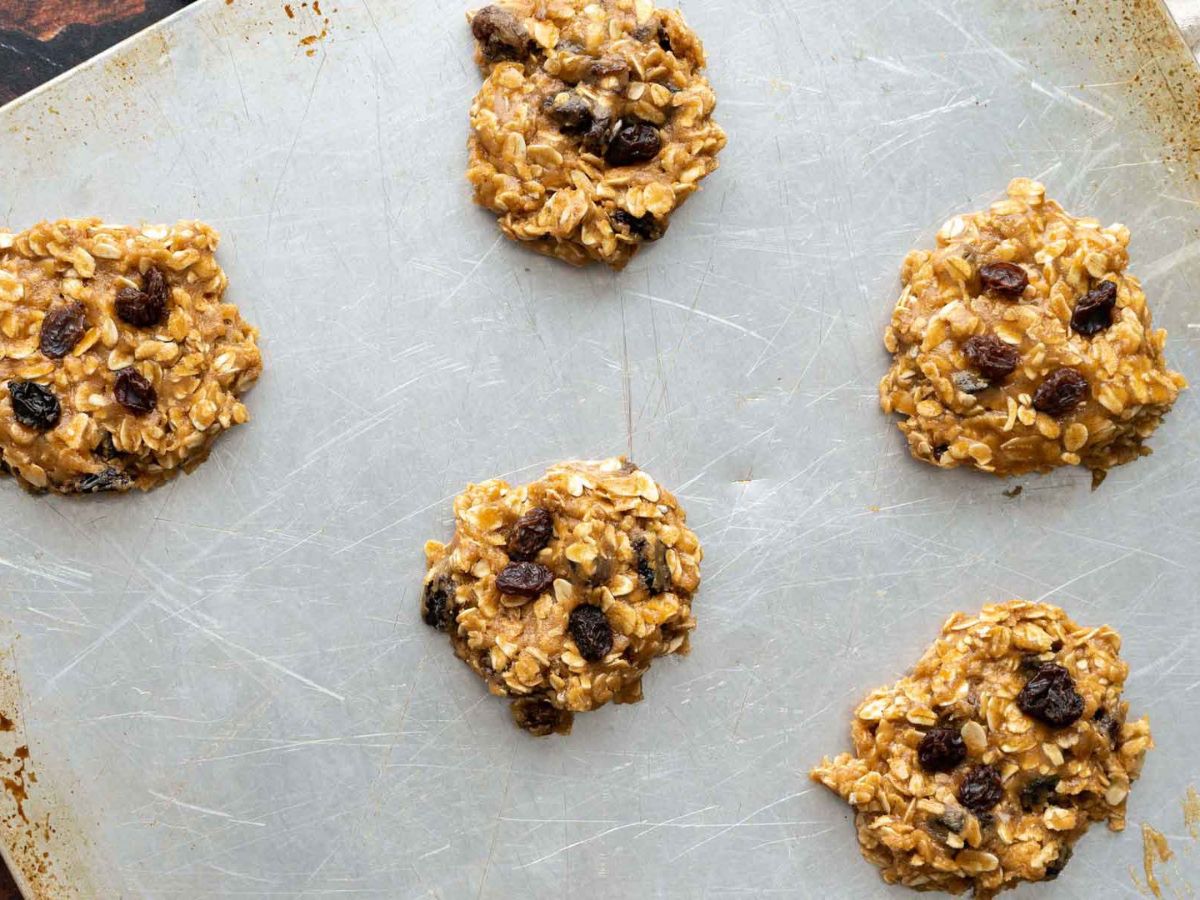 Five unbaked oatmeal raisin cookie dough rounds arranged on a metal baking sheet.