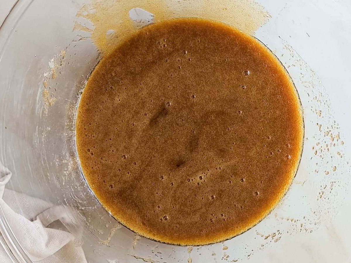 A glass bowl containing a light brown liquid mixture with bubbles on the surface, sitting on a light-colored countertop next to a white cloth.