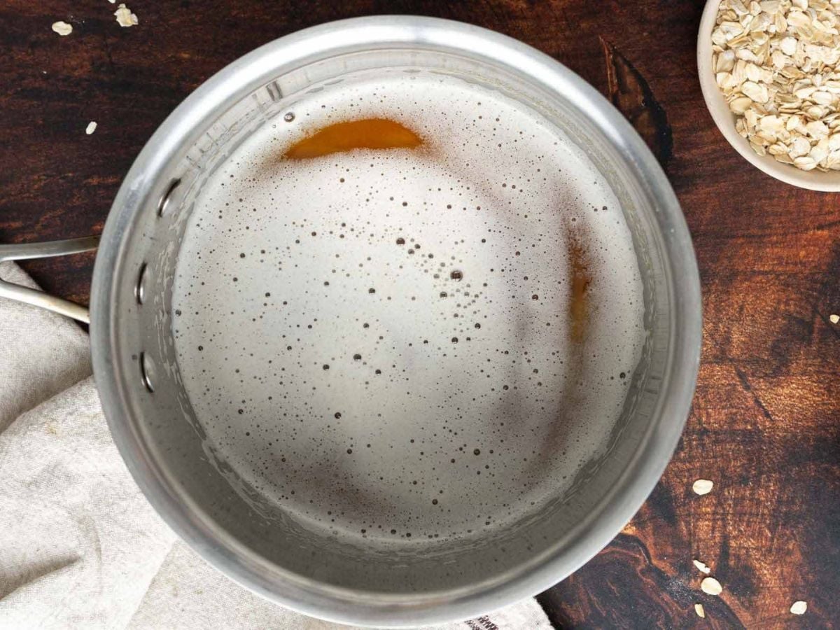 A metal pot filled with foamy liquid sits on a wooden surface next to a bowl of rolled oats and scattered oats.