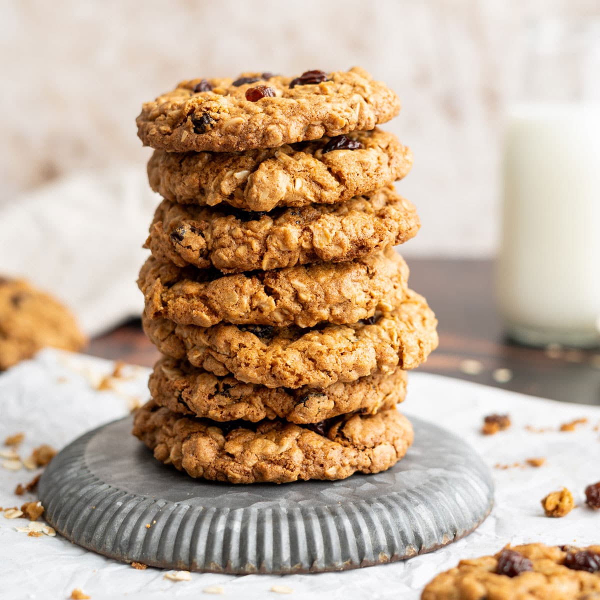 A stack of seven oatmeal raisin cookies sits on a round metal tray, with a glass of milk and scattered cookie crumbs in the background.