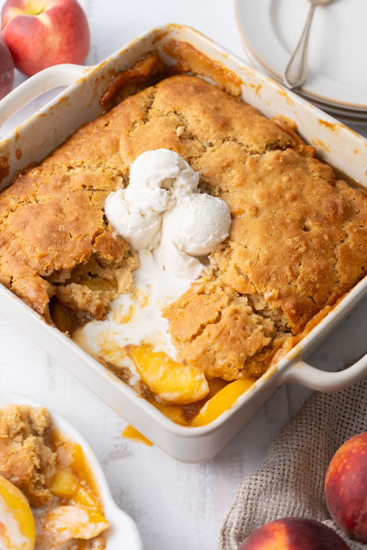 A gluten free peach cobbler in a white dish with a scoop of vanilla ice cream on top, some portions served, surrounded by fresh peaches and plates.