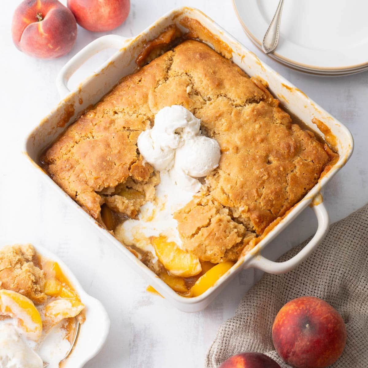 A baked peach cobbler in a square dish, topped with two scoops of vanilla ice cream, with fresh peaches and a plated serving nearby.