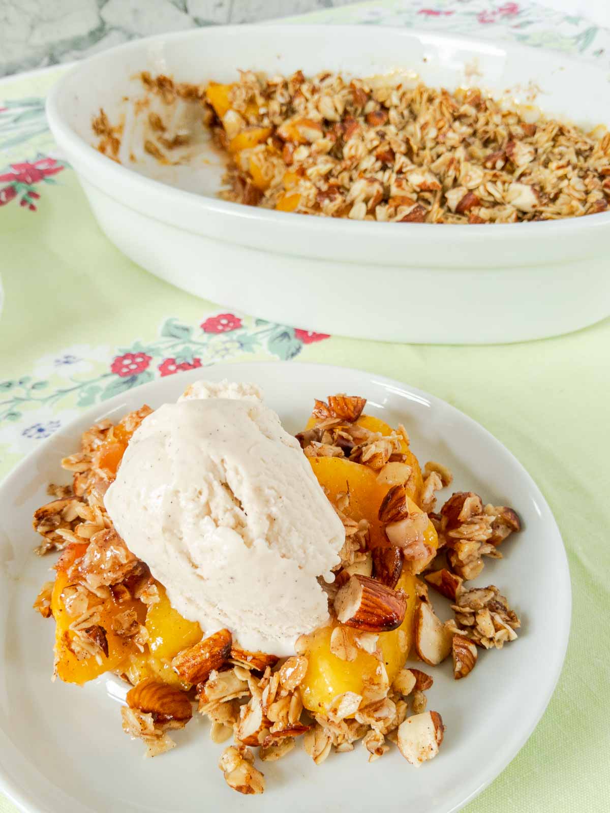 A plate with gluten free peach crisp topped with vanilla ice cream sits in front of a baking dish filled with more peach crisp.