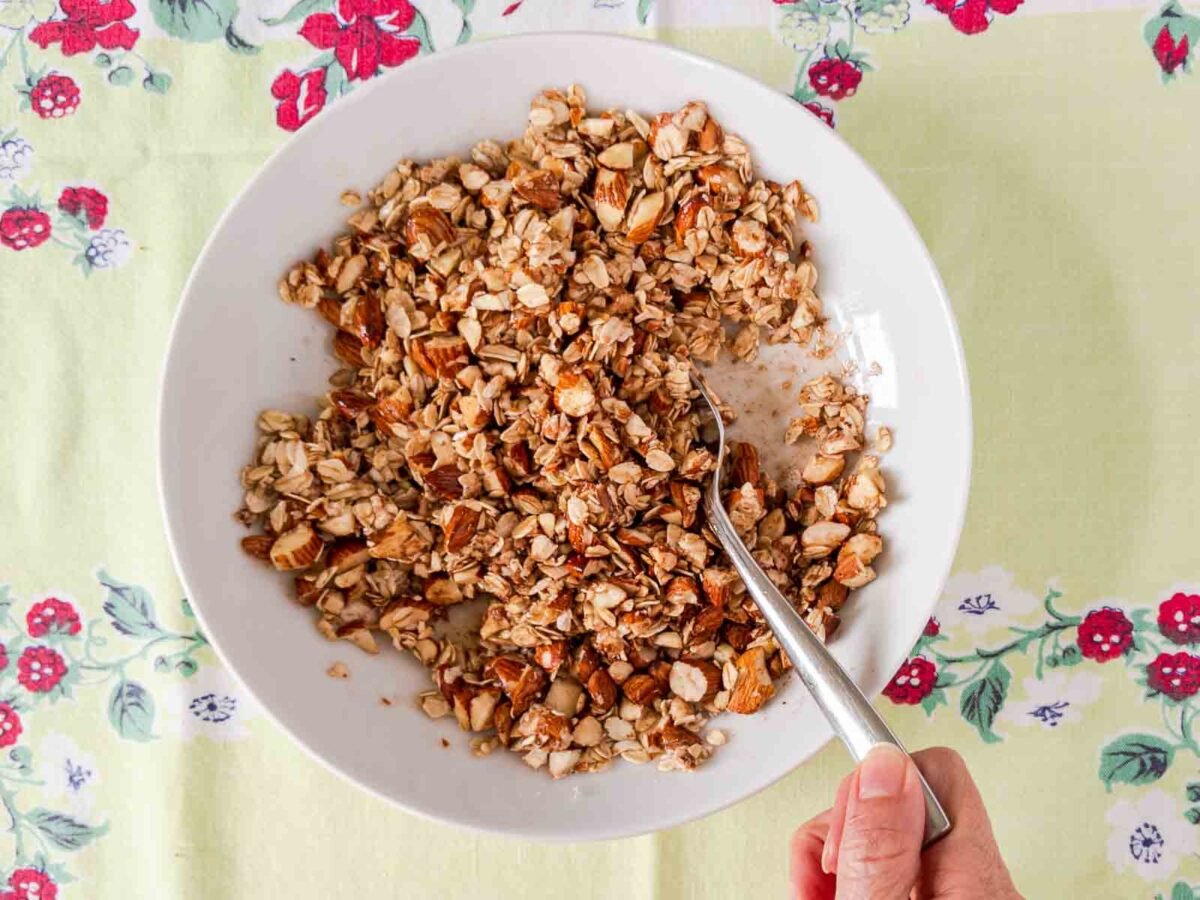 A hand holding a spoon in a bowl of granola with mixed nuts, set on a floral tablecloth, evokes the cozy comfort of gluten free peach crisp.
