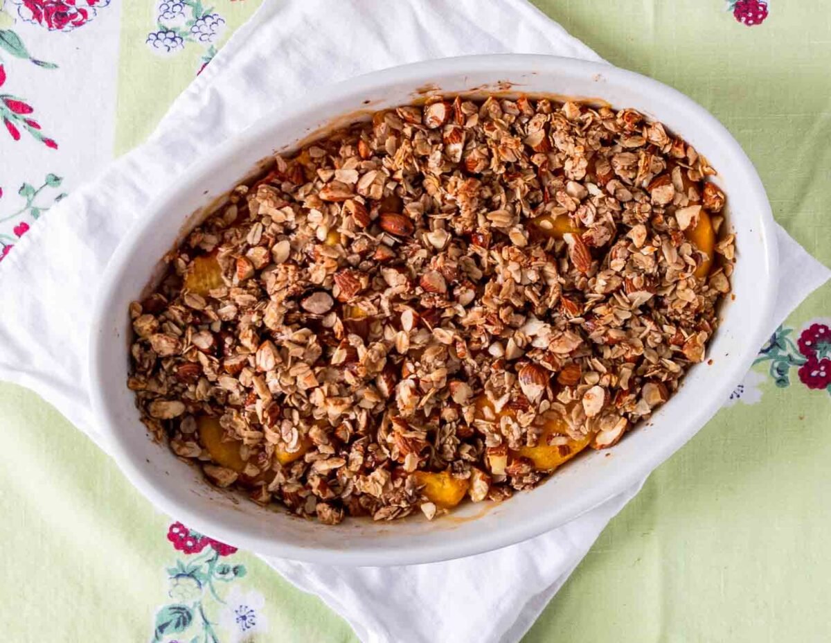 An oval baking dish filled with gluten free peach crisp, topped with oats and chopped nuts, rests on a white cloth atop a green and floral tablecloth.