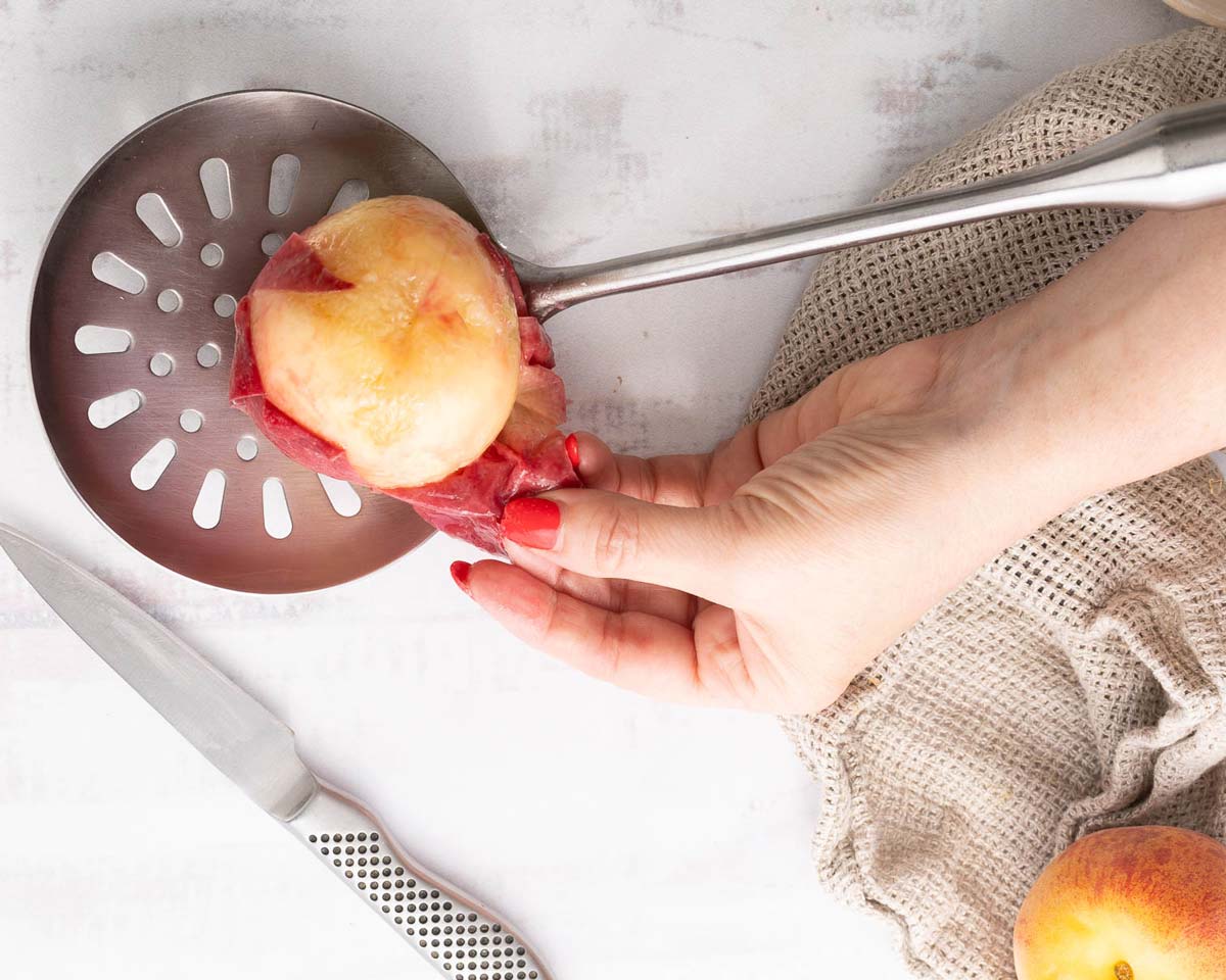 A hand peels the skin off a peach using a slotted spoon, with a knife and a towel nearby on a light surface-perfect prep for making gluten free peach crisp.