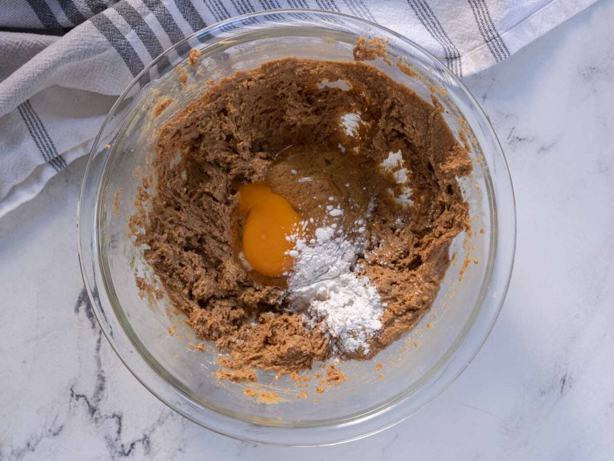 A glass bowl containing brown cookie dough mixture, an egg, and a small amount of flour, on a marble countertop next to a striped kitchen towel.