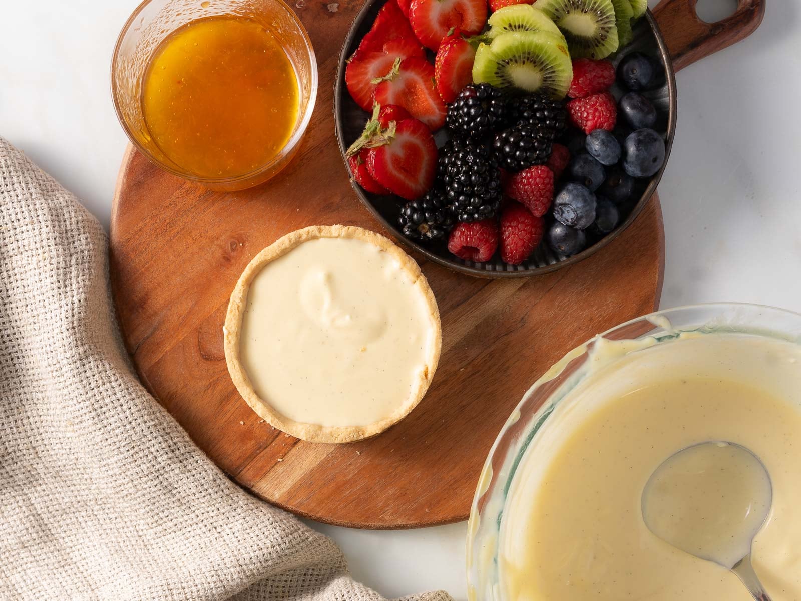 A tart shell filled with cream sits on a wooden board next to a bowl of mixed berries, a glass of orange glaze, a bowl of cream, and a beige cloth.